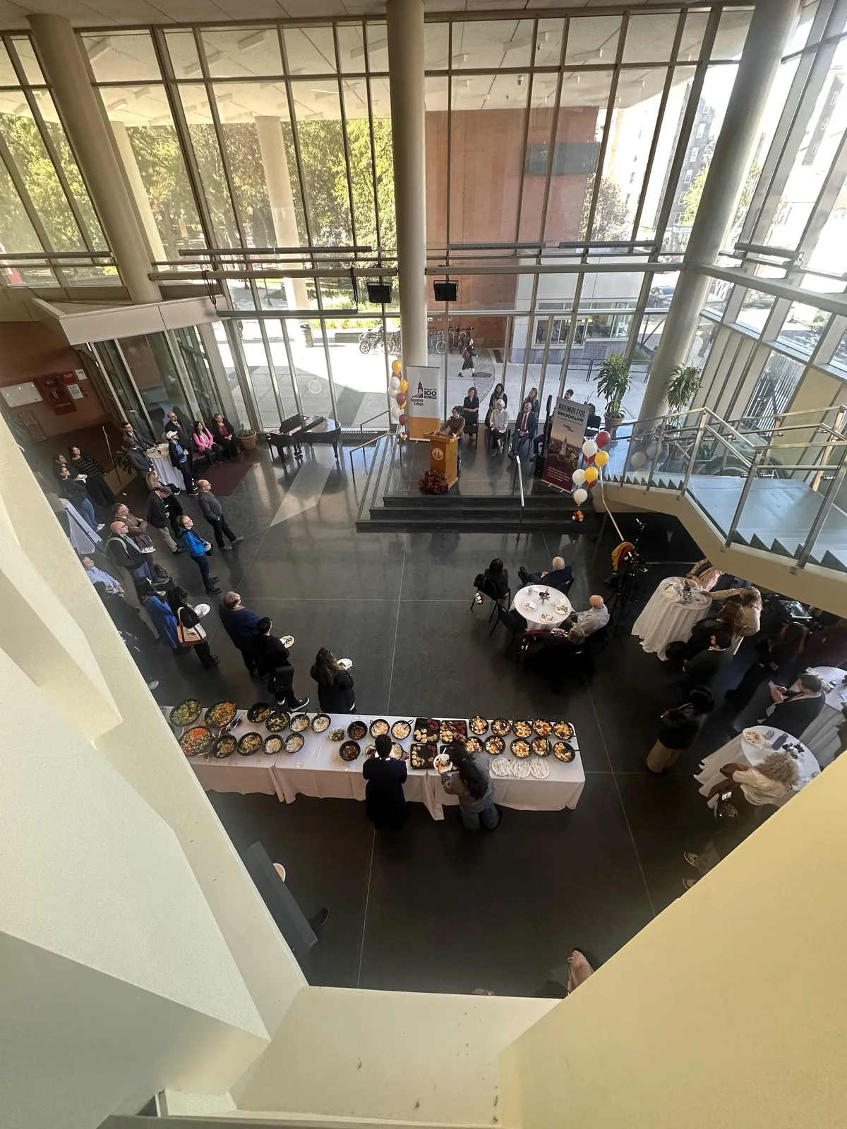 Looking down from a balcony into a glass-walled college atrium during an announcement, with a speaker at a podium, guests seated on stage, a buffet table and a video camera on a tripod by the stairs