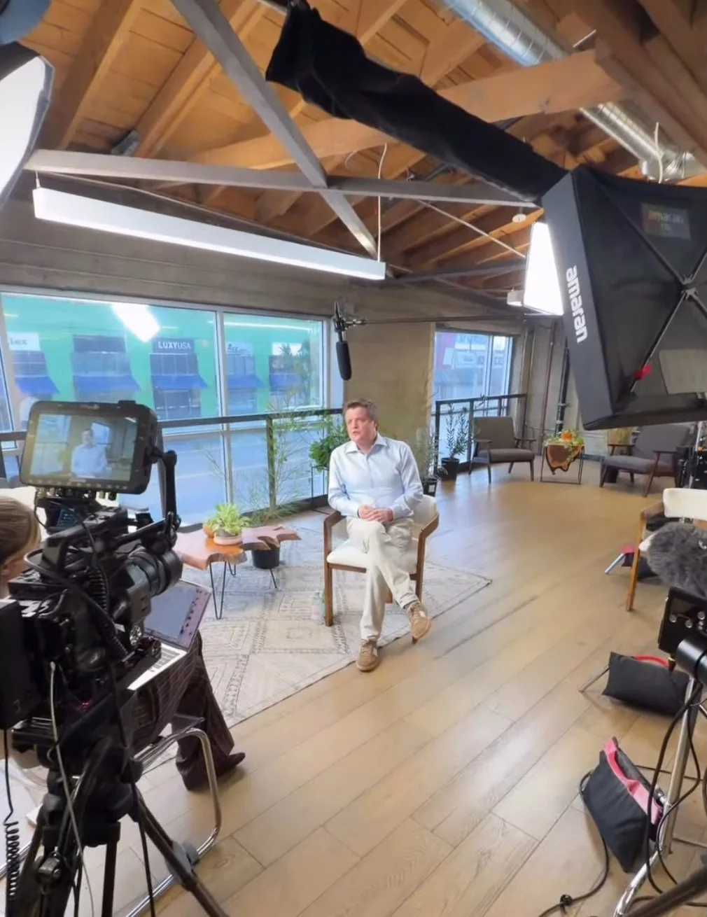 A seated interview in a sunlit loft studio with wood beams, a cinema camera and monitor in the foreground, a large softbox light and a boom microphone overhead