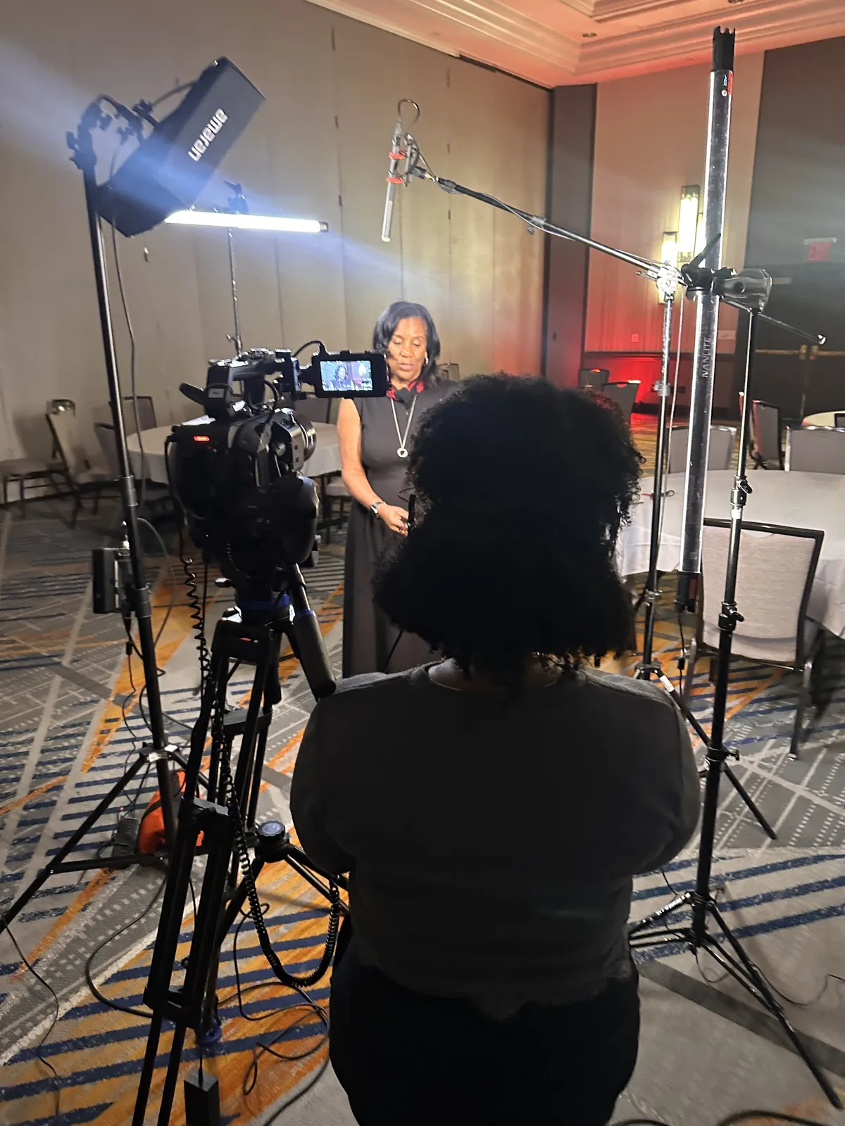 An interview being filmed in a hotel ballroom, with a cinema camera and monitor on a tripod, an LED light, a boom microphone overhead and a producer watching from behind the camera