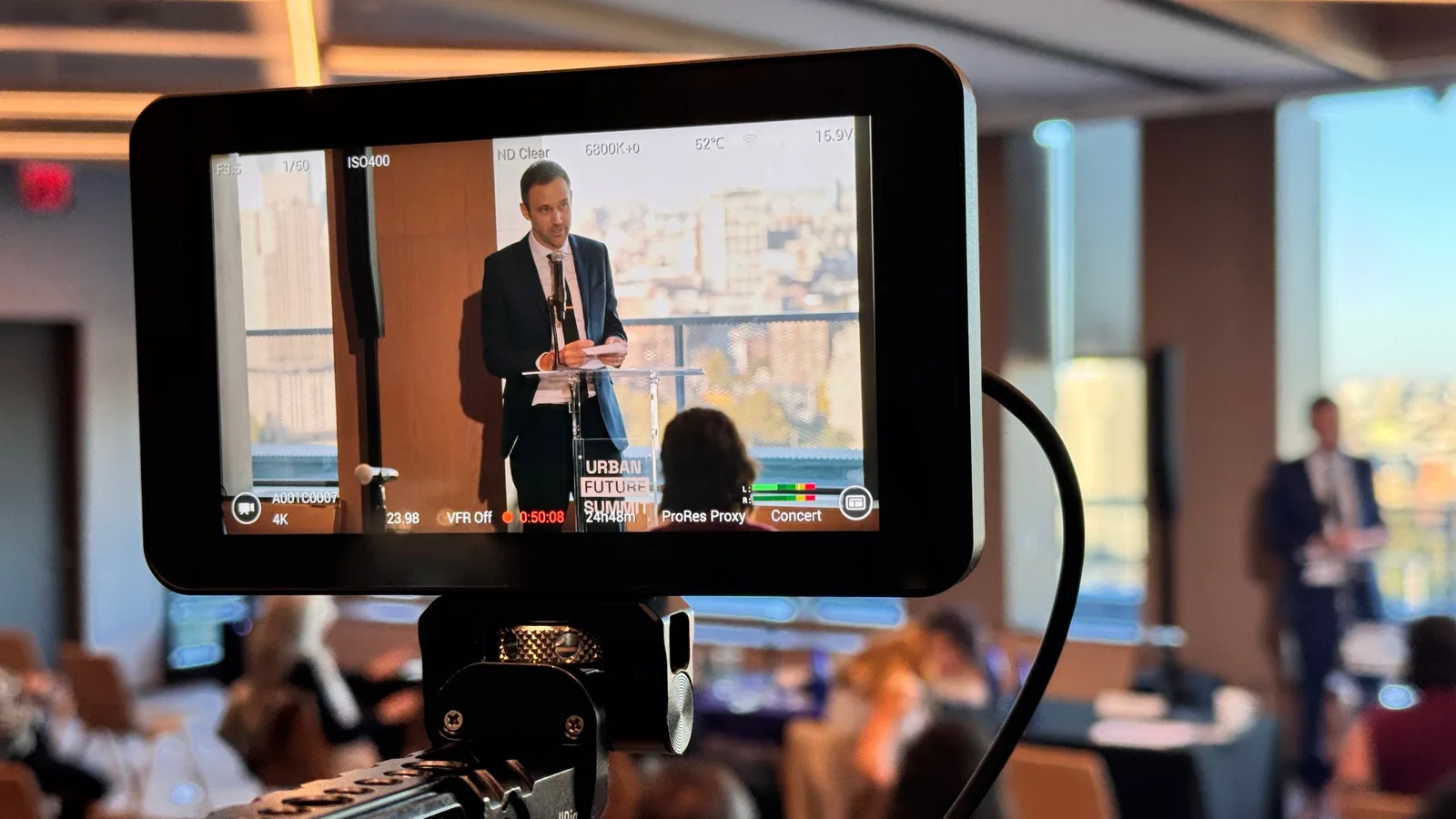 A camera monitor recording a speaker at a podium in 4K, with the live room blurred behind it