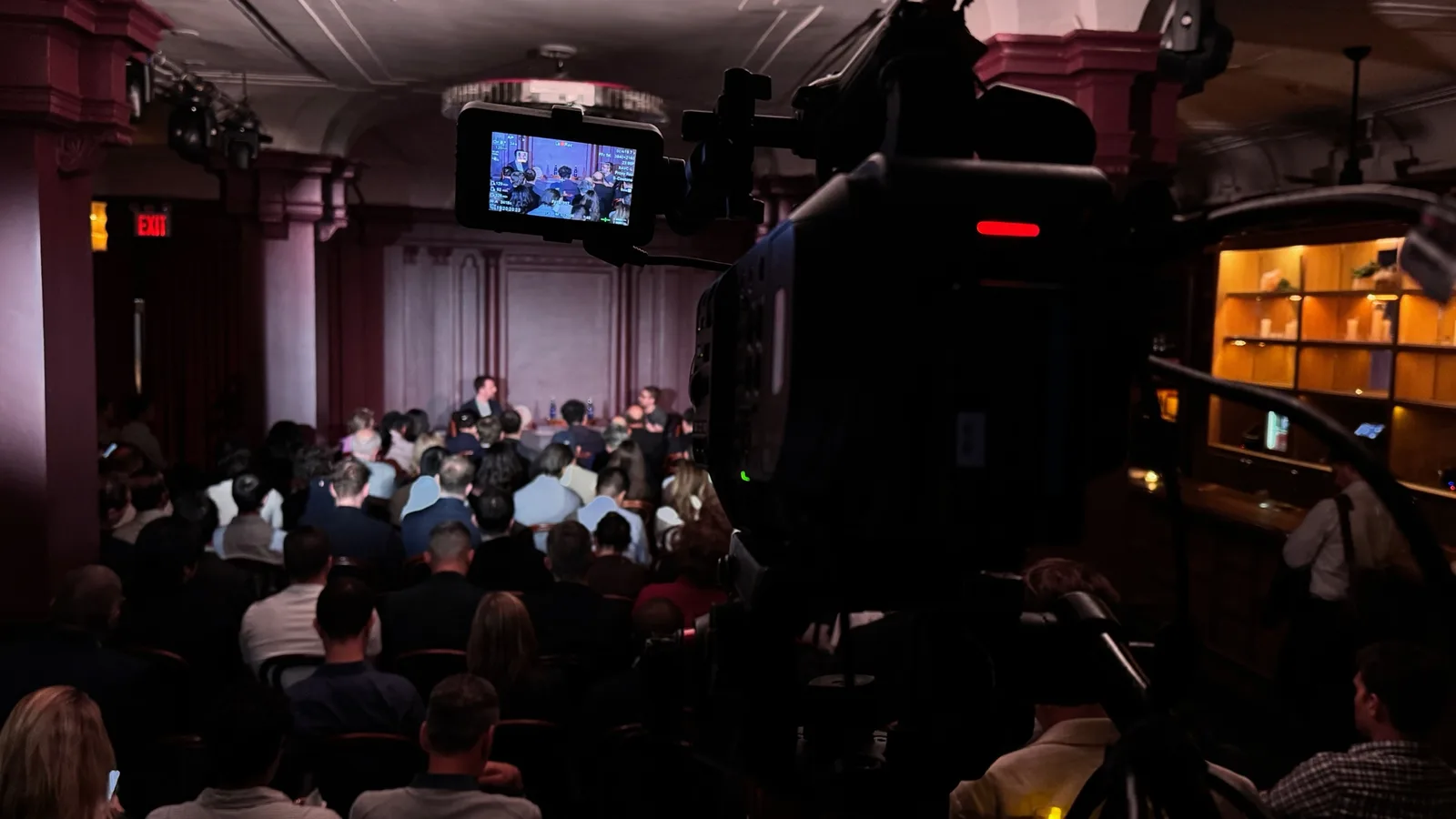 A camera monitor framing a panel discussion in a dim event room