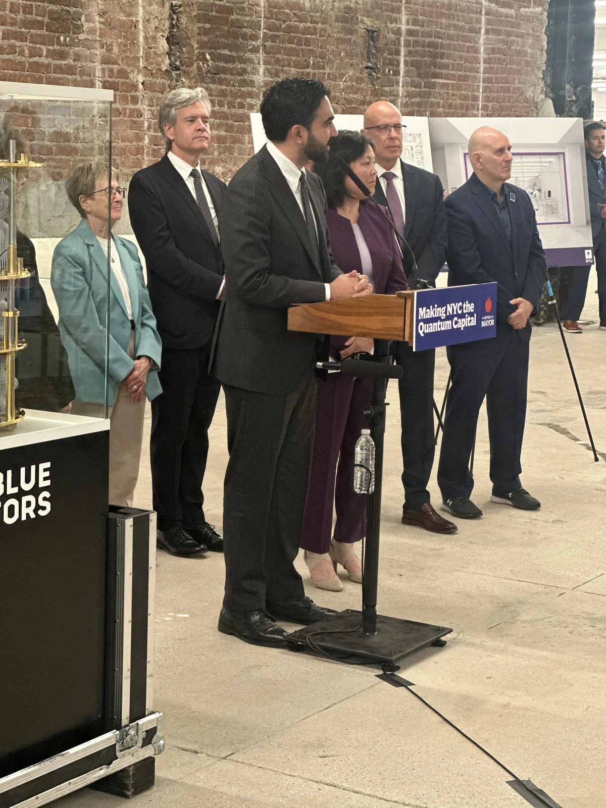 A speaker at a wooden podium with a microphone stand in front of a row of officials