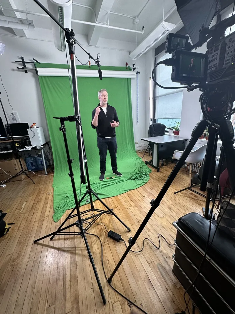 A presenter standing in front of a green screen with a camera and lights in the foreground