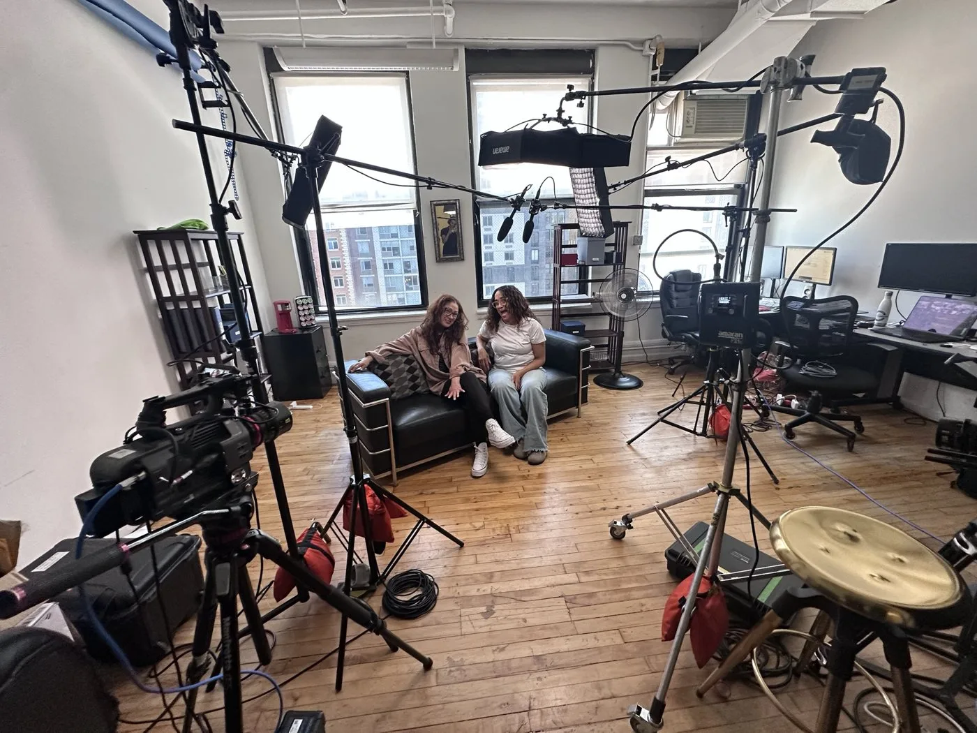 Two guests on a sofa in a sunlit loft studio, with a camera, boom microphone and lights set up for a conversation